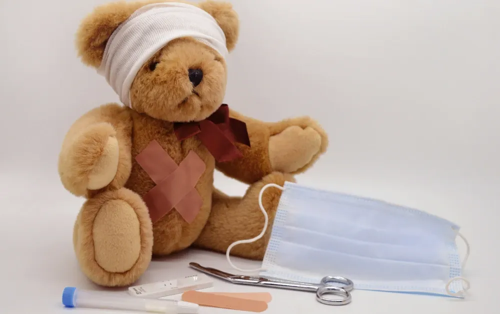 Plišani meda sa flasterima i zavojem na glavi, okružen zaštitnom maskom i medicinskim priborom, simbolizuje dečiju negu.